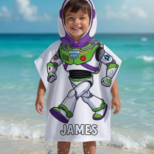 Puede incluir: Toalla de playa blanca con capucha con un dise&ntilde;o de Buzz Lightyear, con detalles verdes, morados y blancos. El nombre "JAMES" est&aacute; impreso en la parte inferior. Un ni&ntilde;o lleva la toalla en la playa.