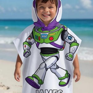Puede incluir: Una toalla de playa con capucha blanca con un dise&ntilde;o de Buzz Lightyear de Toy Story. La toalla tiene una combinaci&oacute;n de colores verde, morado y blanco, con el nombre "JAMES" impreso en negro en la parte inferior. El ni&ntilde;o lleva la toalla en una playa.