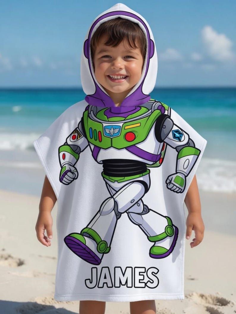 Puede incluir: Una toalla de playa con capucha blanca con un dise&ntilde;o de Buzz Lightyear de Toy Story. La toalla tiene una combinaci&oacute;n de colores verde, morado y blanco, con el nombre "JAMES" impreso en negro en la parte inferior. El ni&ntilde;o lleva la toalla en una playa.