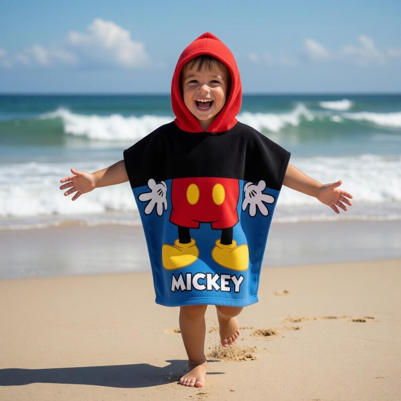 K&ouml;nnte beinhalten: Ein Kind am Strand tr&auml;gt einen Mickey Mouse Poncho mit roter Kapuze. Der Poncho hat einen schwarzen oberen Teil und einen blauen unteren Teil mit Mickeys roter Hose, gelben Schuhen und dem Wort "MICKEY". Das Kind l&auml;chelt mit ausgestreckten Armen.