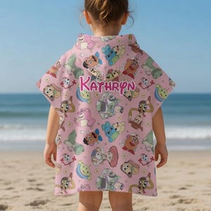 May include: A pink hooded beach towel with a repeating pattern of cartoon dog characters. The name "KATHRYN" is printed in pink script across the back. The child is standing on a sandy beach with the ocean in the background.