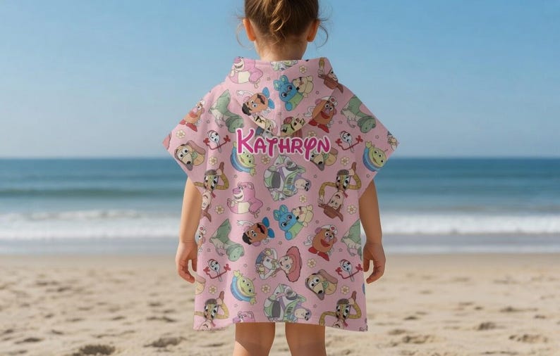 May include: A pink hooded beach towel with a repeating pattern of cartoon dog characters. The name "KATHRYN" is printed in pink script across the back. The child is standing on a sandy beach with the ocean in the background.