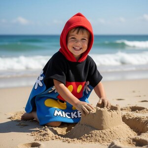 K&ouml;nnte beinhalten: Ein Kind tr&auml;gt einen roten Mickey Mouse Strandtuch-Poncho mit Kapuze und spielt an einem Sandstrand. Der Poncho ist schwarz mit einem roten und gelben Mickey Mouse-Design auf der Vorderseite und blauen Akzenten. Das Wort "MICKEY" ist sichtbar.