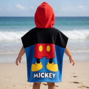 K&ouml;nnte beinhalten: Ein Kapuzen-Strandponcho mit Mickey Mouse-Design. Der Poncho hat eine rote Kapuze, einen schwarzen oberen Teil und einen blauen unteren Teil mit Mickeys roter Hose, gelben Schuhen und dem Namen "MICKEY" in Wei&szlig;. Das Kind steht an einem Sandstrand.