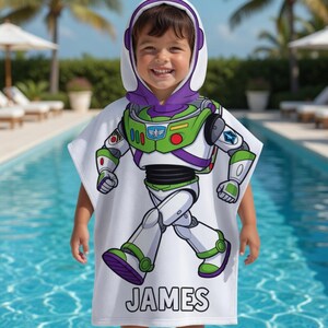 Puede incluir: Una toalla con capucha blanca con un dise&ntilde;o de Buzz Lightyear. La toalla presenta un gr&aacute;fico de cuerpo completo del personaje, con detalles verdes, morados y blancos. El nombre "JAMES" est&aacute; impreso en negro. El ni&ntilde;o est&aacute; junto a una piscina.