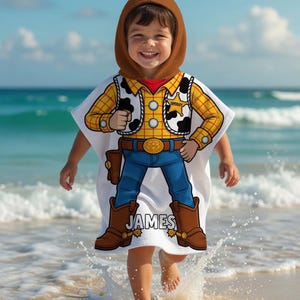May include: A white hooded beach towel featuring a cartoon cowboy design. The towel has a brown hood and a colorful graphic of a cowboy outfit, including a yellow plaid shirt, blue jeans, and brown boots. The name "JAMES" is printed on the towel.