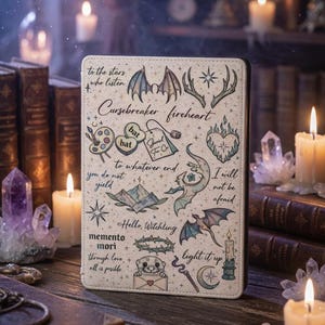 Könnte beinhalten: Eine beige Tablet-Hülle mit verschiedenen skurrilen Illustrationen und Text, darunter Fledermäuse und Geweihe, sowie der Schriftzug "Cursebreaker fireheart". Das Design hat ein mystisches Thema, mit Kerzen und Büchern im Hintergrund.