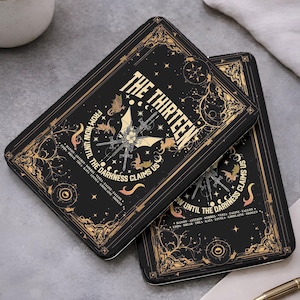 Op de afbeelding: Twee zwarte rechthoekige cases met gouden accenten en de tekst "THE THIRTEEN" in goud. De cases hebben een ontwerp met vleermuizen, zwaarden en de zin "FROM NOW UNTIL THE DARKNESS CLAIMS US".