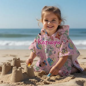 May include: A young child wearing a pink hooded beach towel with cartoon character designs and the name "KATHRYN" in pink lettering. The child is sitting on a sandy beach, smiling, and playing with sand. The ocean is visible in the background.