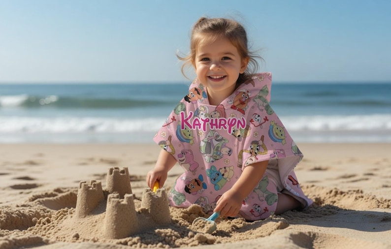 May include: A young child wearing a pink hooded beach towel with cartoon character designs and the name "KATHRYN" in pink lettering. The child is sitting on a sandy beach, smiling, and playing with sand. The ocean is visible in the background.