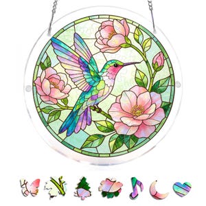 Op de afbeelding: Een rond glazen glas-in-loodraam met een kleurrijke kolibrie met roze, groene en blauwe tinten, die zich in de buurt van roze bloemen bevindt. Het raam wordt opgehangen door een zilveren ketting. Extra decoratieve vormen staan onderaan.
