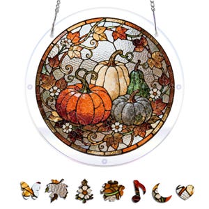May include: A circular stained glass suncatcher featuring a harvest scene with orange, white, and grey pumpkins surrounded by autumn leaves and small white flowers. The piece includes a metal hanging chain and seven distinct puzzle-shaped pieces.