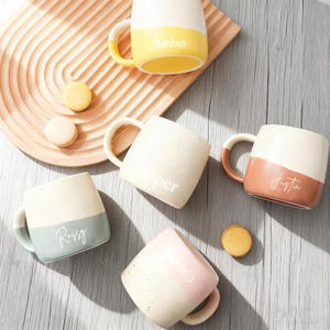 May include: A selection of colour-blocked ceramic mugs, each with a different name in white script. The mugs are in shades of yellow, blue, pink, and brown. They are displayed on a wooden tray and a wooden surface.
