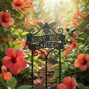 Könnte beinhalten: Schwarzes Metall-Gartenschild mit den Worten "Busy Bee Garden" und einer Bienen-Silhouette. Das Schild befindet sich in einem Garten mit leuchtend orangefarbenen Blumen und einem Kolibri. Das Schild steht auf einem Metallpfahl.