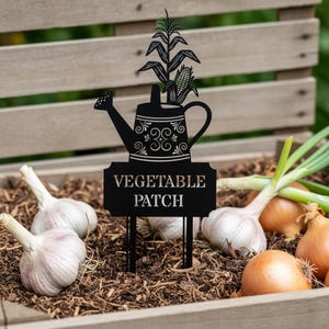 Könnte beinhalten: Schwarzer Metall-Gartenstecker mit Gießkannen-Design und dem Schriftzug "VEGETABLE PATCH". Der Stecker zeigt einen Maisstängel und Maiskolben. Das Bild zeigt auch Knoblauch und Zwiebeln in einem hölzernen Pflanzkasten.
