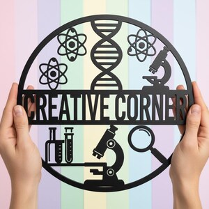 Könnte beinhalten: Ein schwarzes, kreisförmiges Schild mit den ausgeschnittenen Worten "CREATIVE CORNER". Das Schild zeigt wissenschaftliche Ausschnitte, darunter Atome, eine DNA-Strähne, Mikroskope, Reagenzgläser und eine Lupe. Das Schild wird von zwei Händen vor einem pastellfarbenen Hintergrund gehalten.