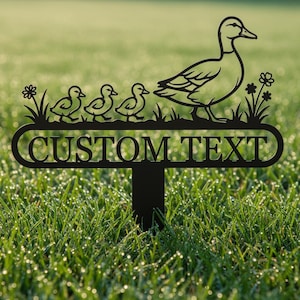 Op de afbeelding: Zwarte metalen tuinpaal met een eend en drie eendenkuikens, met de tekst "CUSTOM TEXT" in een decoratief kader. De paal is ontworpen om in een tuin of gazon te plaatsen, en voegt een persoonlijk tintje toe aan buitenruimtes.