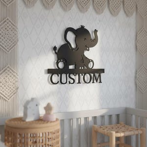 Könnte beinhalten: Schwarze Metall-Wandkunst mit einem Elefanten und dem Wort "CUSTOM" darunter. Der Elefant lächelt und hat seinen Rüssel erhoben. Die Wandkunst wird an einer weißen Wand mit Rautenmuster gezeigt.