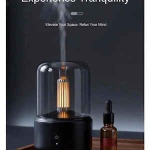 Pu&ograve; includere: Un moderno diffusore di aromi con la scritta "Experience Tranquility". La base nera supporta una copertura cilindrica trasparente. Una piccola bottiglia di olio essenziale &egrave; accanto al diffusore. L'immagine trasmette un senso di relax e benessere.