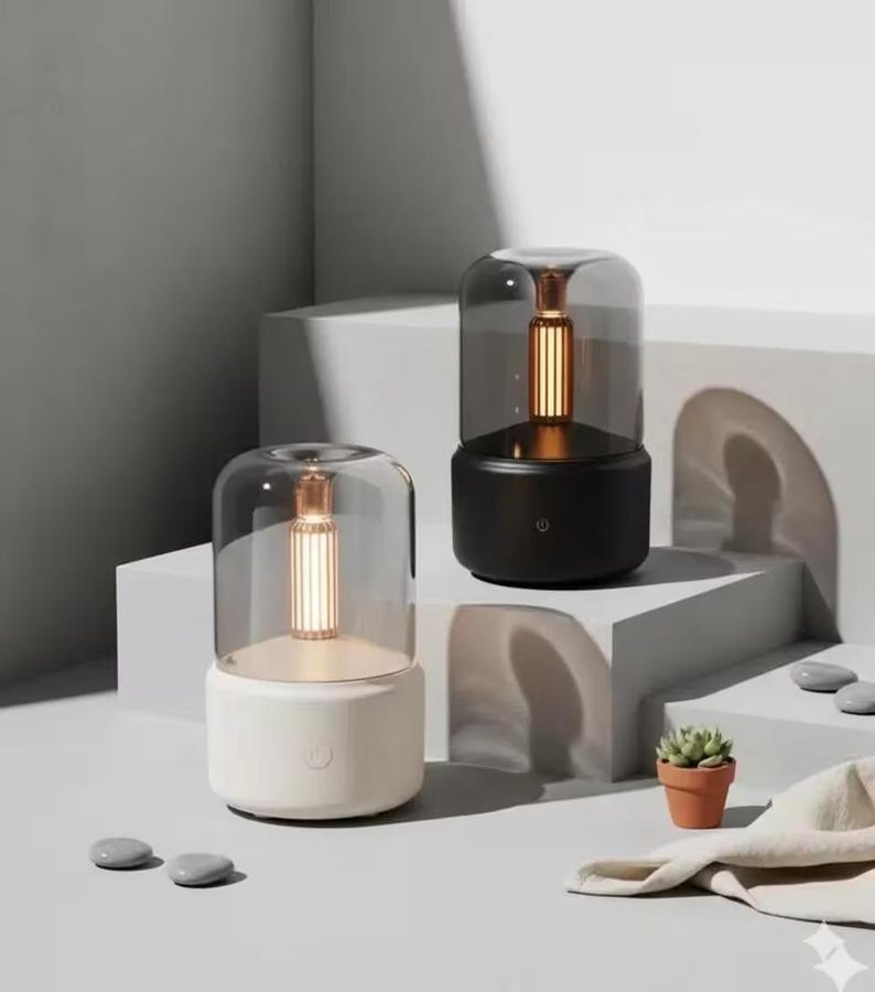 Pu&ograve; includere: Due moderne lampade da tavolo con paralume cilindrico in vetro e luce calda. Una lampada &egrave; bianca, l'altra nera. Sono posizionate su una superficie bianca, con una piccola pianta in vaso e pietre.