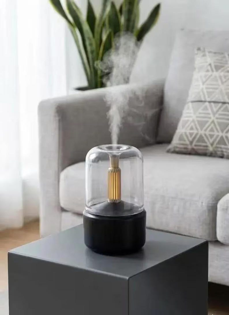 Pu&ograve; includere: Un diffusore di oli essenziali nero e trasparente con un elemento centrale color oro. Il diffusore emette un flusso di vapore. &Egrave; posizionato su un tavolo a forma di cubo grigio scuro, con un divano grigio e una pianta sullo sfondo.