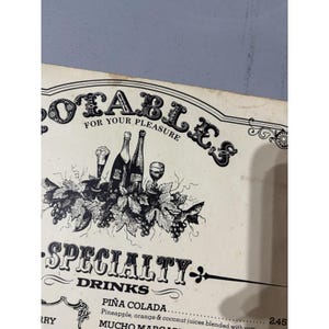 Puede incluir: Un menú de estilo vintage con la palabra "DOTABLES" en letras ornamentadas. Debajo, se muestra el texto "FOR YOUR PLEASURE". El menú presenta ilustraciones de botellas de vino, copas y vides, con las palabras "SPECIALTY DRINKS" debajo.