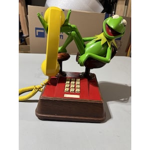 Könnte beinhalten: Ein neuartiges Telefon mit einer grünen Kermit-der-Frosch-Figur, die auf einem braunen Stuhl liegt. Das Telefon hat eine rote Tastatur, einen gelben Hörer und einen braunen Sockel. Die Kermit-Figur ist grün mit rotem Mund und weißen Augen.