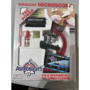 May include: Vintage Tasco microscope set for young astronauts. The red and black microscope is packaged with accessories and a super-lite lamp. The box features the text "Young Astronauts" and "View Specimens 750 Times Closer."