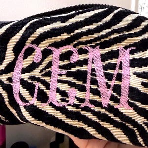 Op de afbeelding: Een tasje met rits voorzien van een zwart en beige zebrastreeppatroon met een textuurafwerking. De voorkant van het tasje is geborduurd met de letters GEM in lichtroze draad, in een decoratieve schreefletterstijl in het midden.