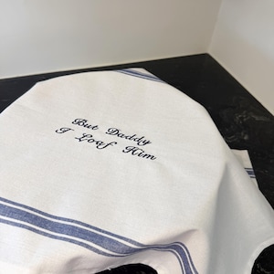 May include: White and blue striped kitchen towel with the embroidered text "But Daddy I Loaf Him". The towel is made of a soft, light-colored fabric and has two blue stripes running along the edges.