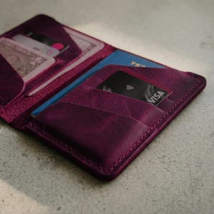 May include: A close-up of a deep purple leather wallet, open to reveal a blue passport, cash, and credit cards. The wallet has multiple pockets and is stitched with a matching thread. The VISA card is visible in one of the pockets.