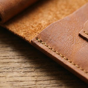 K&ouml;nnte beinhalten: Nahaufnahme einer handgefertigten, offenen Ledergeldb&ouml;rse. Die Geldb&ouml;rse hat eine warme braune Farbe mit sichtbaren N&auml;hten aus kontrastierendem hellbraunem Faden. Das Leder hat eine strukturierte Oberfl&auml;che und die Kanten sind sauber verarbeitet.