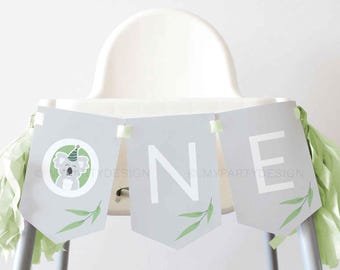 May include: A gray banner with the word "ONE" in white letters, decorated with a koala illustration and green leaf accents. The banner is adorned with green tassels and is displayed on a high chair. The website address "MYPARTYDESIGN.COM" is printed at the bottom.