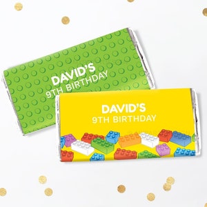 Può includere: Due involucri di barrette di cioccolato a tema Lego. Un involucro è verde con pois bianchi e dice "David's 9th Birthday". L'altro involucro è giallo con un motivo di mattoncini Lego colorati e dice "David's 9th Birthday".