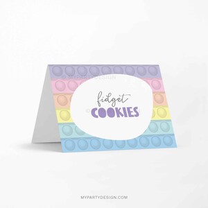 Może przedstawiać: Złożona karta z kolorowym wzorem pop-it. Karta ma pastelową tęczową kolorystykę ze słowami "fidget COOKIES" w odręcznej czcionce w owalu. Karta jest biała w środku.
