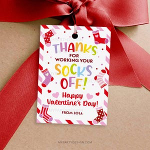 May include: A Valentine's Day gift tag with a red ribbon. The tag has a white background with red and pink accents, featuring colorful socks and the text "THANKS FOR WORKING YOUR SOCKS OFF! Happy Valentine's Day!" and "FROM LOLA".