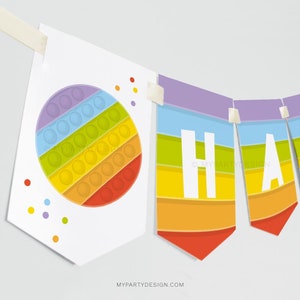 Może przedstawiać: Baner w kolorze tęczy z napisem "Happy" białymi literami. Baner składa się z pojedynczych flag z wzorem tęczy i wzorem pop it.