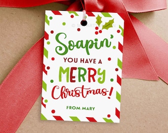 Op de afbeelding: Een feestelijk cadeaulabel met een rode strik. Het label heeft een witte achtergrond met groene en rode accenten. De tekst "Soapin' You Have a Merry Christmas!" is geschreven in groen en rood. Het label is ondertekend met "From Mary".