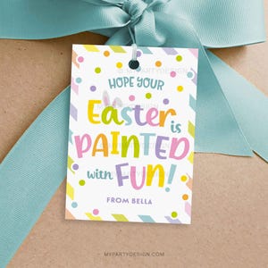 May include: A colourful Easter gift tag with the message "Hope your Easter is painted with FUN!" The tag is decorated with pastel polka dots and geometric shapes. A light blue ribbon is tied around the gift.