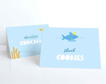 May include: Two light blue table cards with text and illustrations. One card says "chocolate CUPCAKE" and the other says "Shark COOKIES" with a cartoon shark wearing a party hat. The cards have an underwater theme.