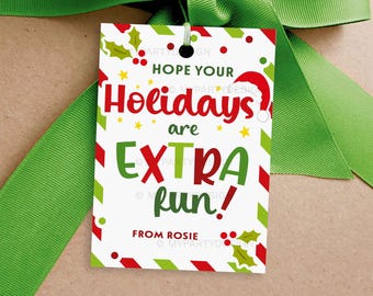 May include: A white gift tag with red and green accents, a green ribbon, and the text "Hope Your Holidays are EXTRA fun!" in red and green. The tag includes a Santa hat and holly illustrations. The tag is tied to a brown package.