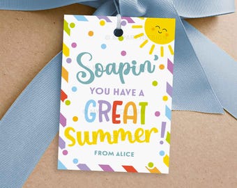 May include: A white gift tag with the text "Soapin' You Have a Great Summer!" in colorful lettering. The tag features a smiling sun graphic and colorful confetti. A light blue ribbon is tied around the gift.