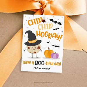 May include: A Halloween-themed gift tag with a cartoon cookie wearing a witch hat, pumpkins, and bats. The tag reads "Chip Chip Hooray!" and "Have a BOO-tiful day!" with a name at the bottom. The tag is tied with an orange ribbon.