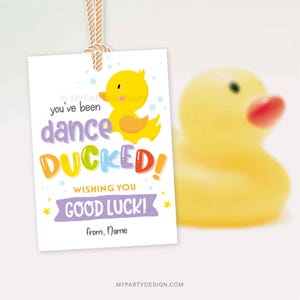 Könnte beinhalten: Ein weißes Geschenk-Etikett mit einer gelben Quietscheente und dem Text "you've been dance DUCKED! Wishing you good luck!" in bunten Buchstaben. Eine gelbe Quietscheente mit rotem Schnabel im Hintergrund.