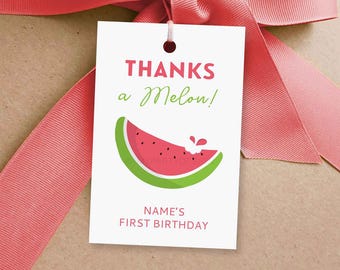 Peut inclure: Une étiquette cadeau blanche avec le texte "THANKS a Melon!" en rose et vert, avec un dessin de tranche de pastèque. L'étiquette est attachée avec un ruban rose et porte la mention "NAME'S FIRST BIRTHDAY".