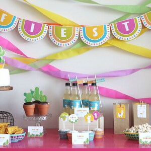 Pu&ograve; includere: Una tavola da festa a tema fiesta colorata con una torta bianca decorata con un lama e cactus, cupcake con topper a forma di lama, bottiglie di limonata, una ciotola di popcorn e un cartello che dice "Prendi pure uno".