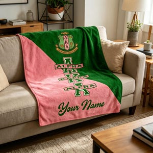 Op de afbeelding: Een groene en roze deken op een beige bank. De deken toont de Griekse letters Alpha Kappa Alpha, een wapen en de woorden "Your Name" in een scriptlettertype. De deken is gemaakt van een zacht, pluche materiaal.