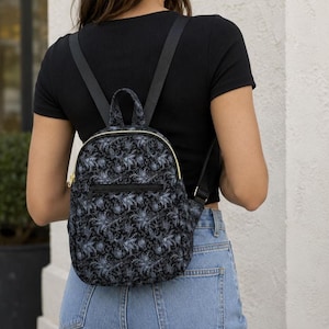 May include: A small backpack featuring a dark black and grey floral damask pattern. The bag includes a top handle, adjustable black shoulder straps, a gold-toned zipper closure, and a horizontal zippered front pocket for additional storage.