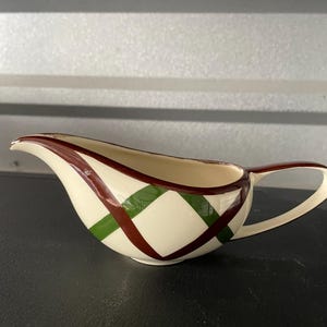 May include: Cream-colored gravy boat with a handle and spout. The boat features a crisscross pattern of dark red and green lines. The gravy boat is made of ceramic.