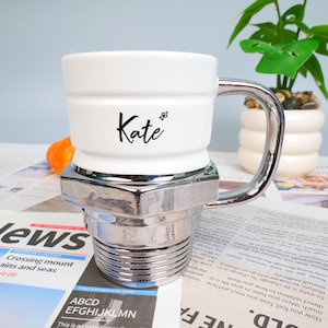 Puede incluir: Taza de cerámica blanca con asa y base plateadas, con el nombre "Kate" en escritura negra. La taza tiene un diseño único, que se asemeja a un perno y una tuerca. Una pequeña fruta naranja y una planta en una maceta blanca están en el fondo.